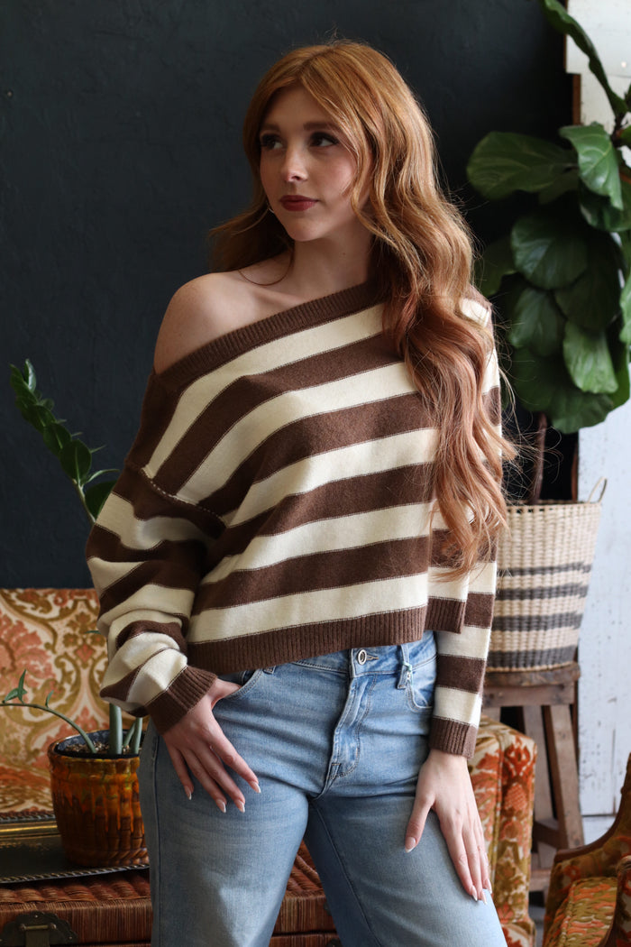 A woman with long wavy red hair models a brown and cream striped off-the-shoulder knit sweater paired with light-wash blue jeans in a cozy indoor setting with plants and a patterned sofa.